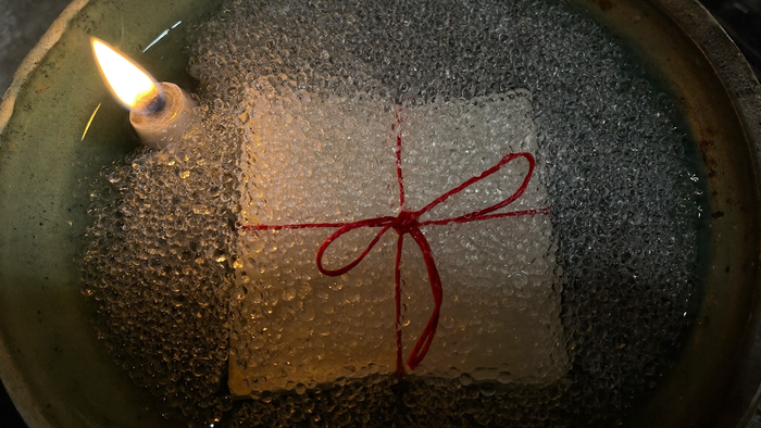 Folded paper tied with red thread frozen under frosted ice in a dark stoneware bowl beside a lit candle