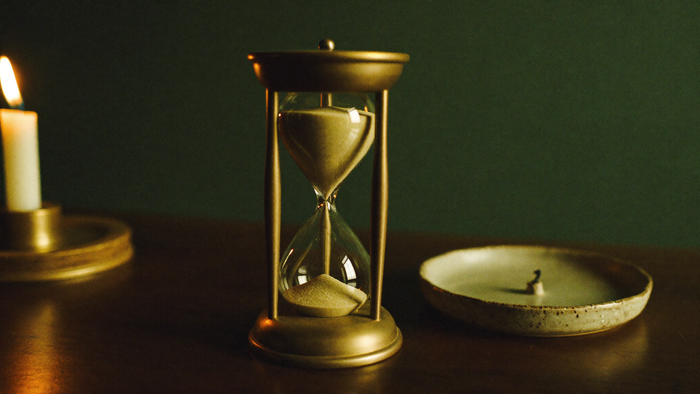 Brass hourglass with the sand running through, on a dark wood table beside a shallow ceramic dish holding a burnt-down candle stub