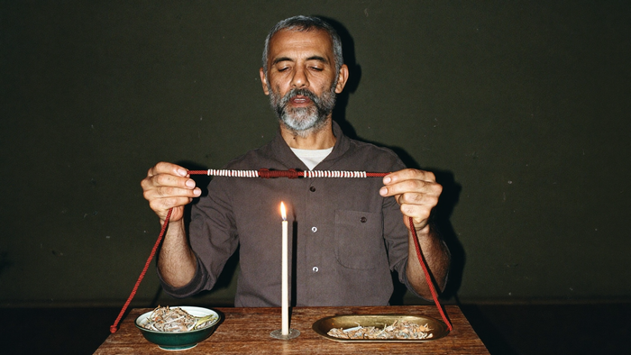 Bearded man with eyes closed holding a knotted red cord above a burning white candle at a small wooden altar