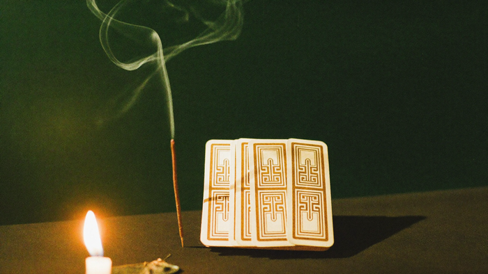 A stack of cards with patterned backs beside a burning incense stick, smoke rising in warm candlelight