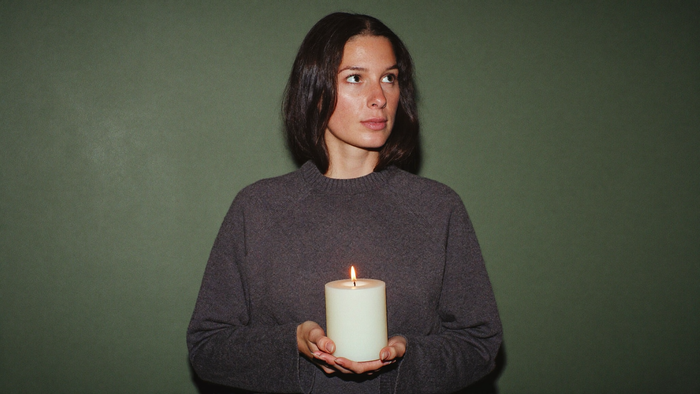 Woman in a gray sweater holding a lit white pillar candle in both hands against a dark green wall