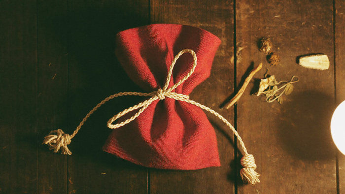 Small red flannel mojo bag tied shut with cord on dark wood, dried roots and herbs lying beside it