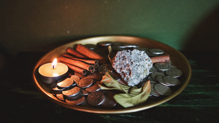 Brass money bowl filled with coins, cinnamon sticks, bay leaves and a raw stone beside a burning candle on dark stone