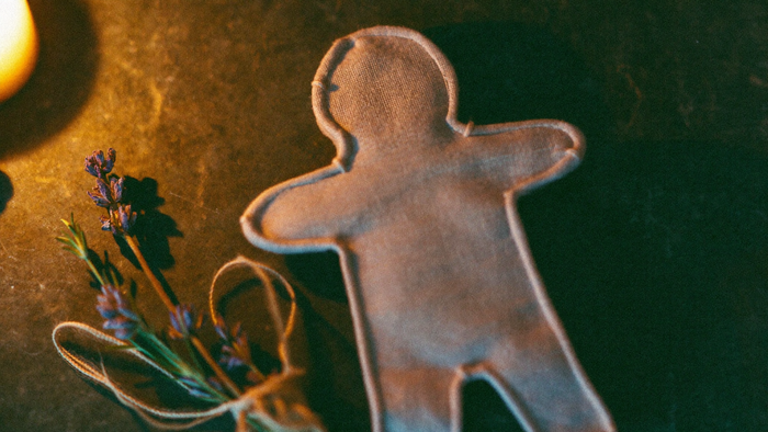 Small linen poppet figure with visible running stitches lying on dark stone beside dried lavender and natural thread