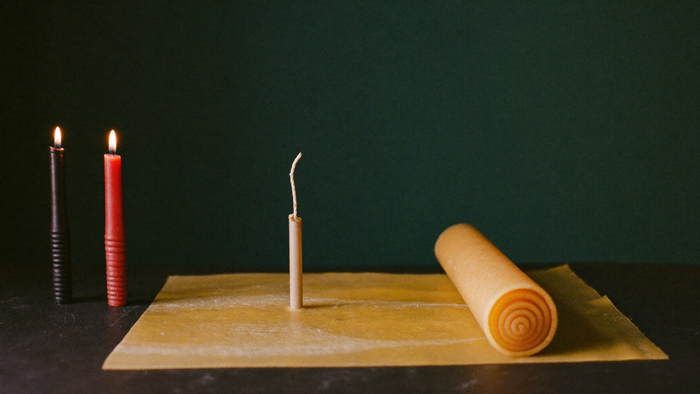 Beeswax sheet with a partly rolled candle and a bare wick beside two burning hand-rolled spell candles on dark stone