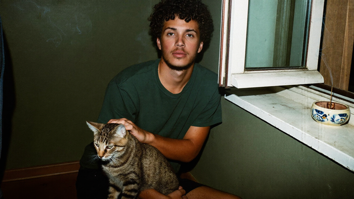 A man sits with a tabby cat on his lap beside an open window while an incense stick smokes on the sill