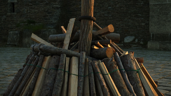 Unlit pyre: a thick wooden stake ringed with a dark band, split logs and brushwood stacked around it on cobblestones