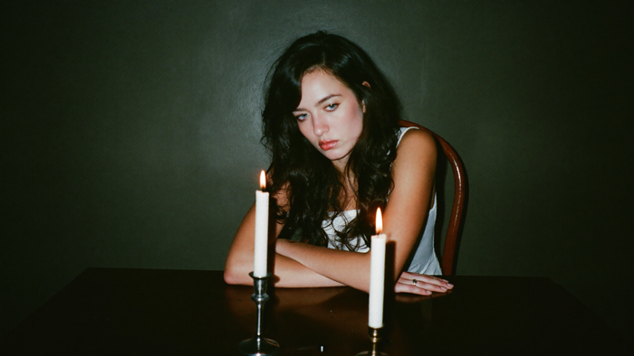 A woman sitting alone at a dark table behind two burning candles, lit by hard direct flash.