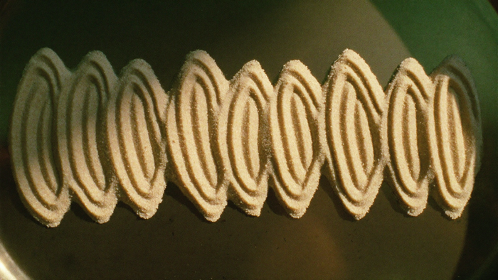 Fine pale sand on a dark metal plate settled into a symmetric standing-wave pattern of ridges under raking light
