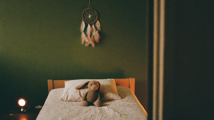 A child's bed with a worn stuffed rabbit on the pillow, a feathered dreamcatcher on the wall and a small night light