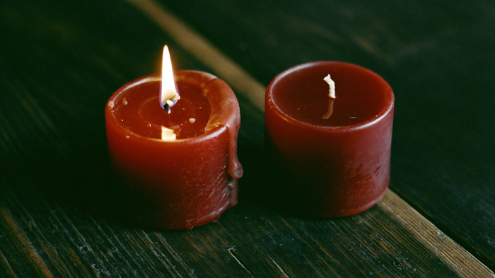 Two dark red candles on weathered wood, the left one burning and the right one still unlit