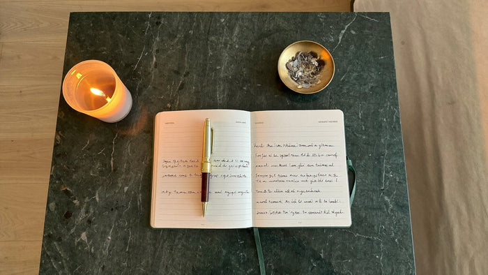 An open dream journal with a pen beside a lit candle and a small brass bowl of dried herbs