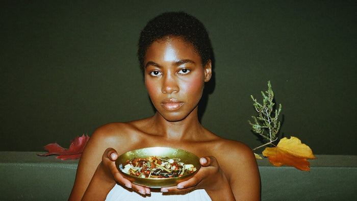 A woman holding a brass bowl of dried herbs and resin beside autumn leaves and a juniper sprig