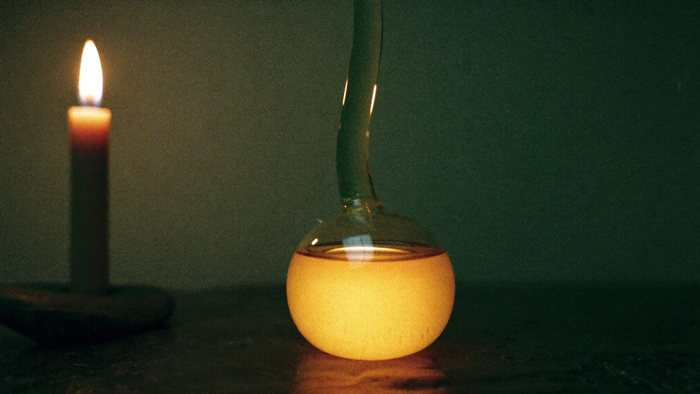 Round glass alchemical flask with glowing amber liquid on dark stone, a lit candle to the left