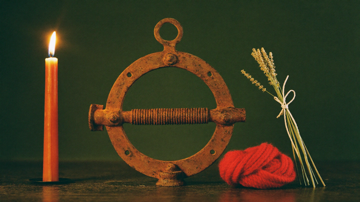 A rusted iron ring, a coil of red wool cord and dried yarrow stand beside a lit candle on dark green.