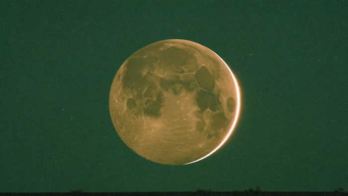 The moon almost fully in shadow with a thin bright rim of sunlight along one edge above a dark horizon