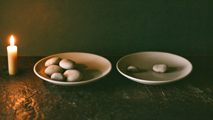 Two shallow dishes on dark stone, pebbles heaped in one and a few moved into the other beside a burning candle