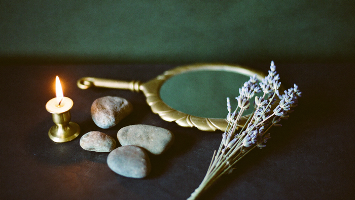 Brass hand mirror beside a lit candle, three smooth stones and dried lavender on a dark stone surface