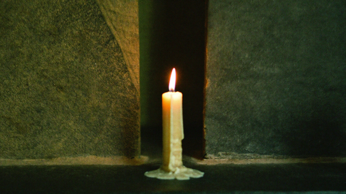 A single lit candle on dark stone before a narrow gap between two slabs with warm light spilling through