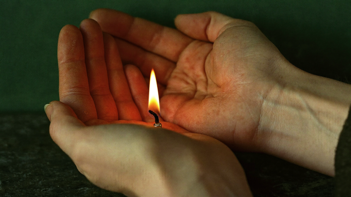 Two hands cupped around a small candle flame on dark stone, warm light falling on the palms