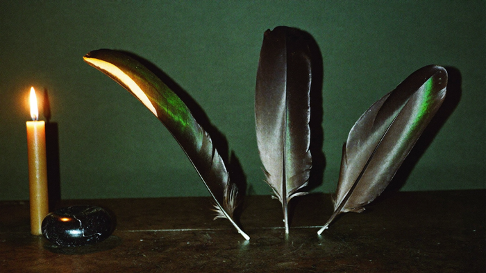 Three black raven feathers and a piece of obsidian on dark stone beside a burning candle