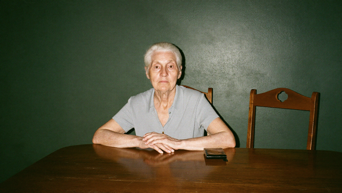 Older woman sitting alone at a dark wooden table, hands clasped in front of her, an empty chair beside her
