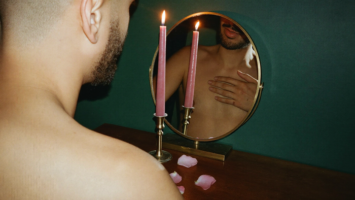 Man with his hand on his heart looking at his reflection in a brass mirror behind two pink candles and rose petals