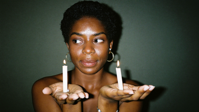 Woman holding two identical lit white candles in her open palms against a dark green wall