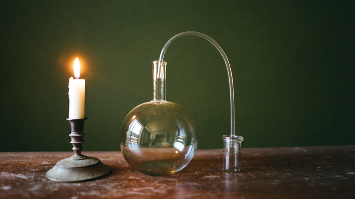 Round-bottomed glass flask with a curved arm running into a small receiving jar beside a lit candle