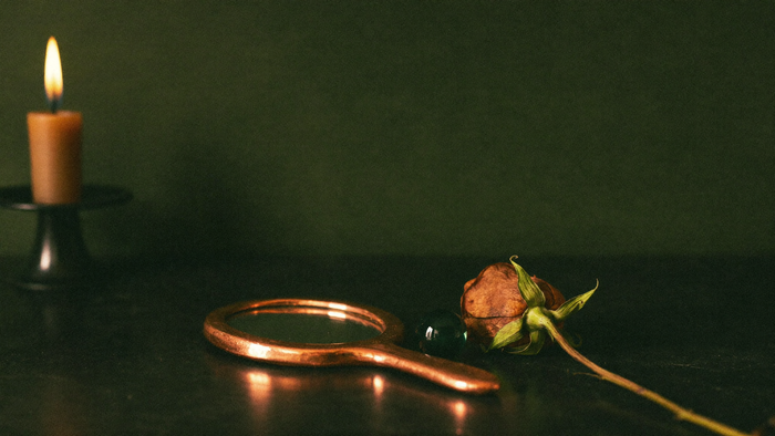 Copper hand mirror lying beside a dried rose and a small green glass bead in warm candlelight