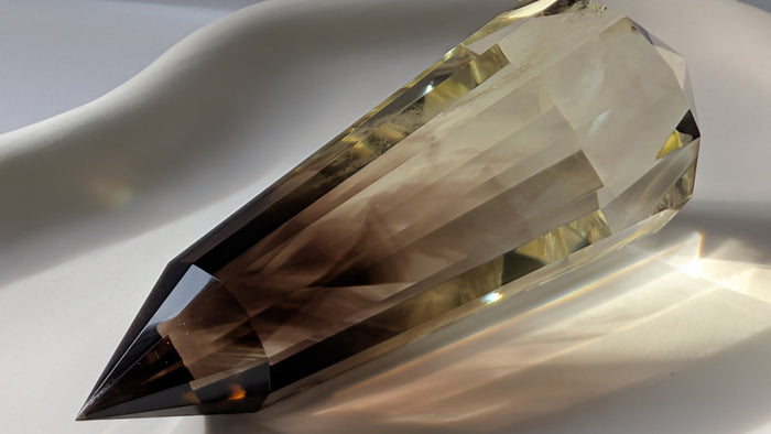 A long faceted double-terminated smoky quartz wand lying on a white surface, light refracting through its cut faces