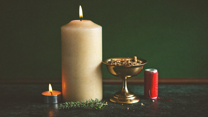 Ivory pillar candle, a brass bowl of smoldering herbs and a spool of red thread on a dark altar stone