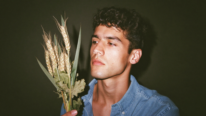 A young man holding a small bundle of dried wheat and oak leaves against a dark green wall