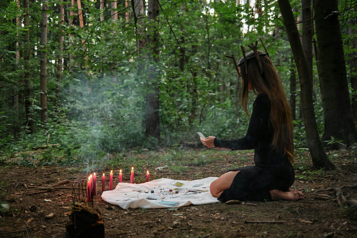 Practitioner kneeling on a white cloth in a forest clearing beside a row of lit red candles, cards and rising smoke