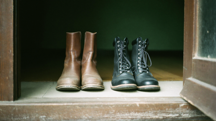 Two pairs of boots side by side on a wooden threshold, one worn brown leather pair and one dark laced pair, soft light
