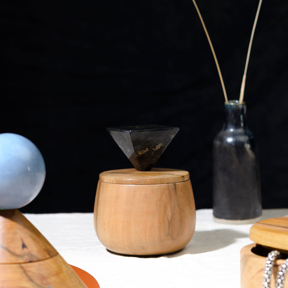 Round pale wood jewel box topped by a faceted dark stone pyramid finial, standing on white cloth against a black background between a pale blue sphere on a wooden stand and a dark speckled bottle holding reed sticks.