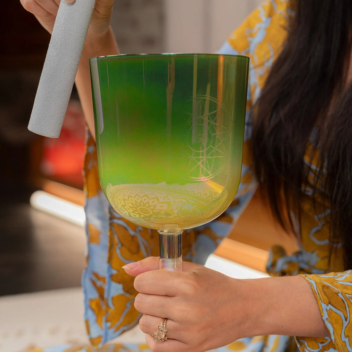 The green iridescent chalice bowl held by its clear stem, etched with a star-and-compass emblem on the side and a mandala band near the base, suede mallet raised above the rim.