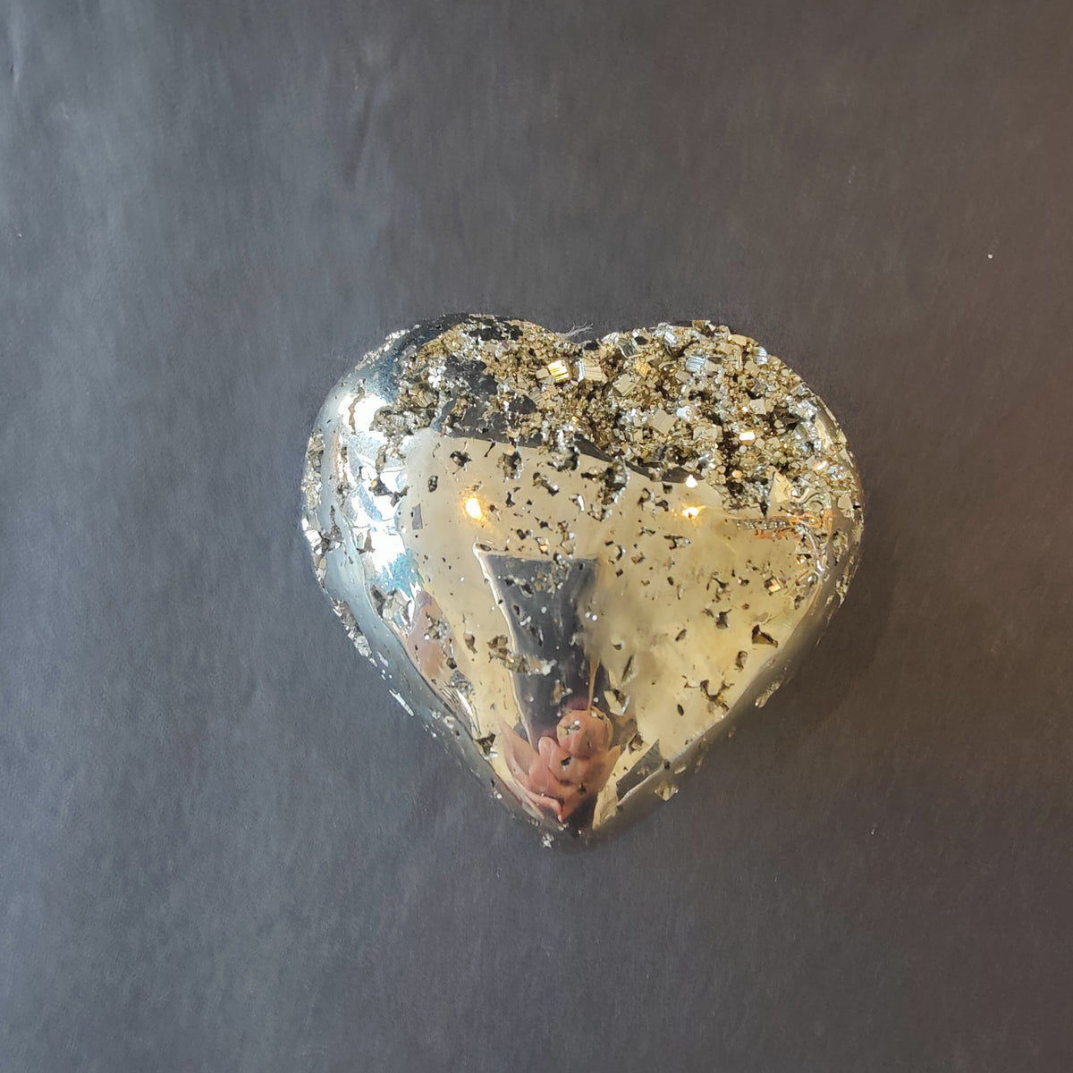 Polished pyrite heart resting on a gray surface, glossy gold face flecked with raw crystal