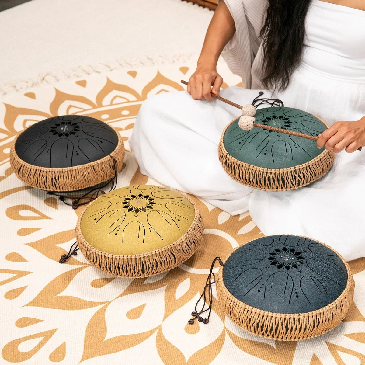 Four steel tongue drums in black, teal, gold and slate blue stand on a mandala rug; a woman in white plays the teal one with two mallets.