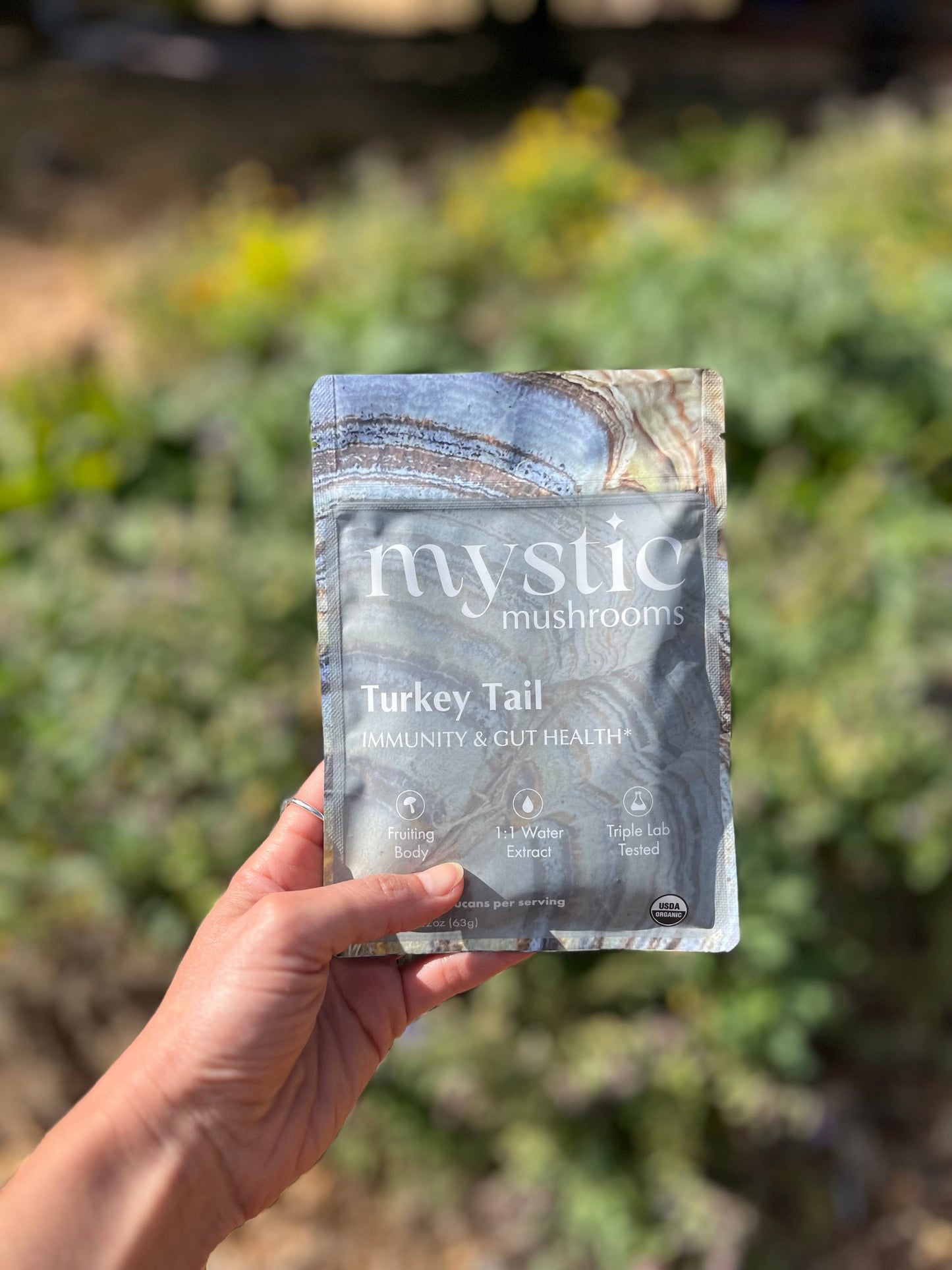 A hand holding the Mystic Mushrooms Turkey Tail pouch outdoors in front of green shrubs, showing the banded turkey tail mushroom artwork