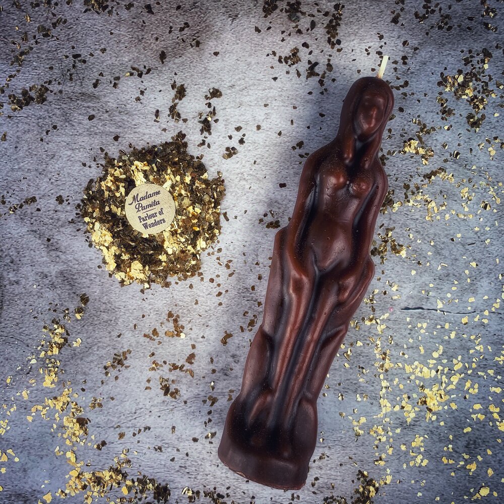 Dark brown beeswax female figure candle on gray cloth with a ring of crushed herbs