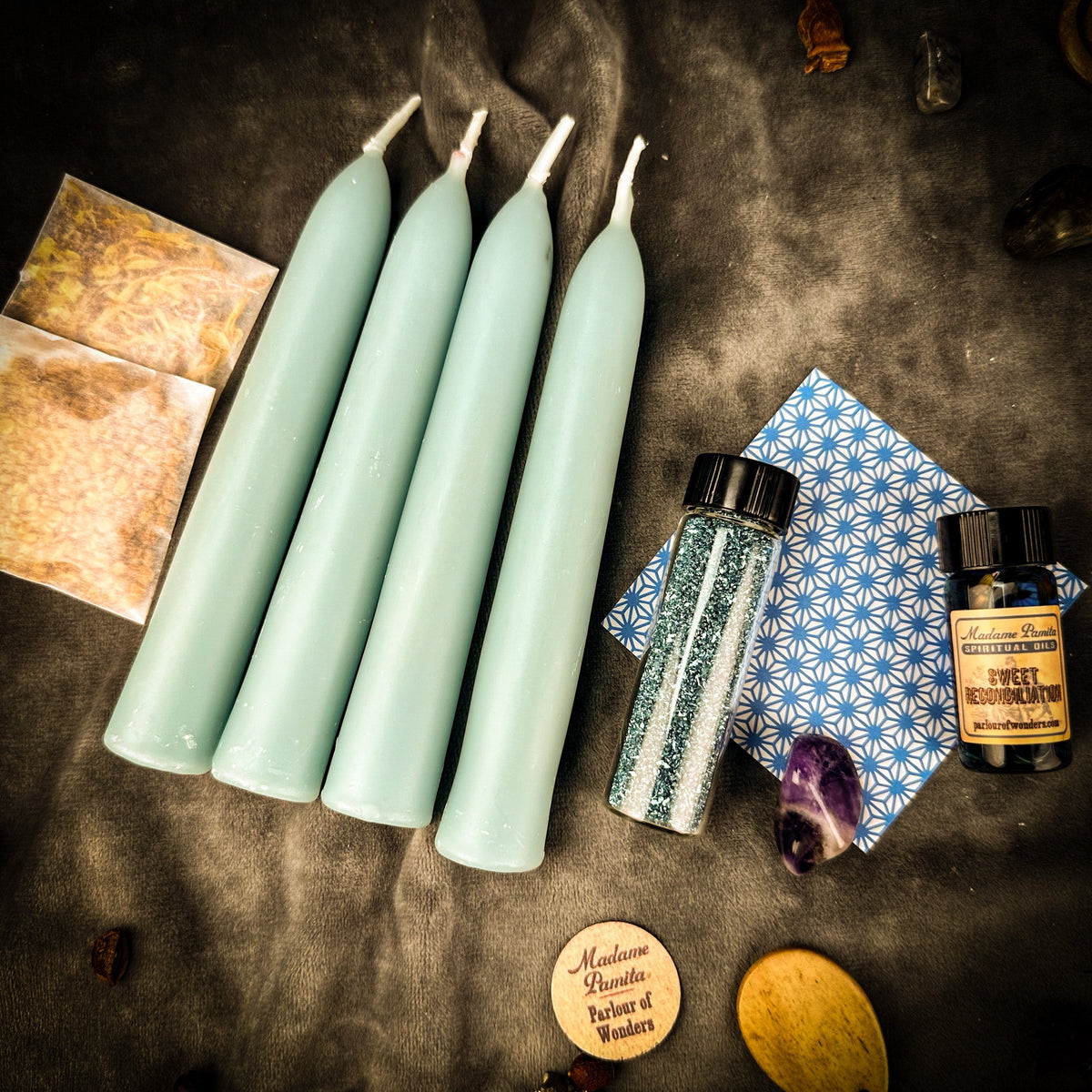 The same Reconciliation pack on dark gray cloth under warm light, which reads the blue tapers a soft sage; a wooden spoon and dried pods lie at the edge.