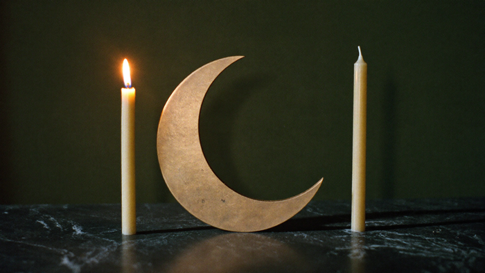 Golden crescent moon between two beeswax taper candles, one of them lit, on dark marble for the 10/10 new moon