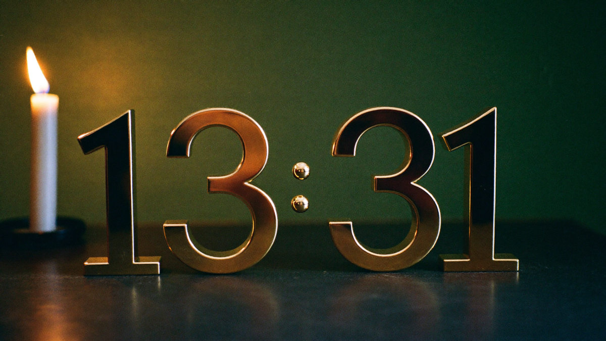 Large brass numerals 13:31 on dark stone in front of a deep green wall, with a lit white candle on the left