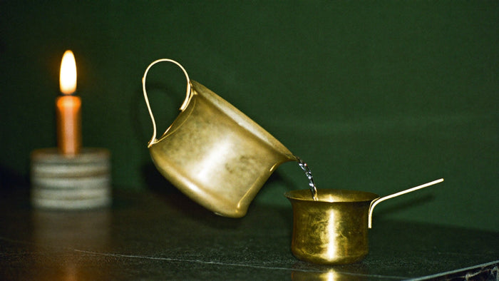 Brass pitcher pouring a thin stream of water into a small brass cup, a candle glowing behind, the image of Temperance