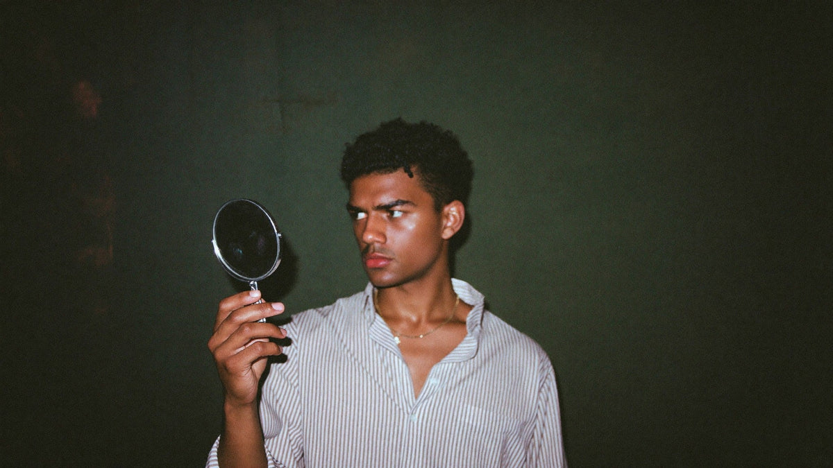 Young man in a striped shirt holding a small round hand mirror up in a dark green room, looking into it late at night