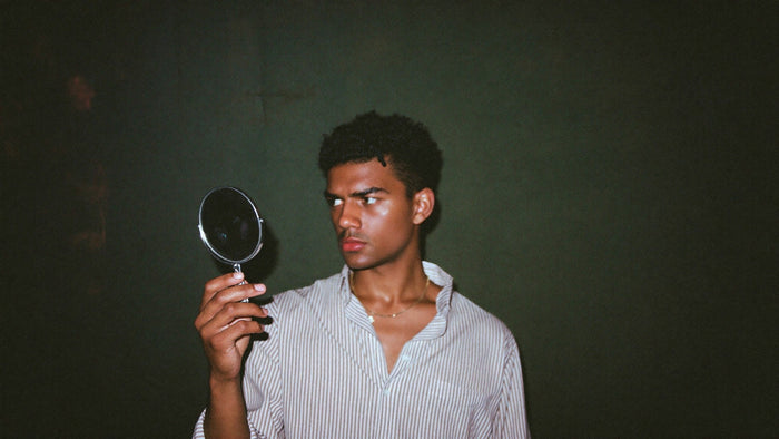 Young man in a striped shirt holding a small round hand mirror up in a dark green room, looking into it late at night