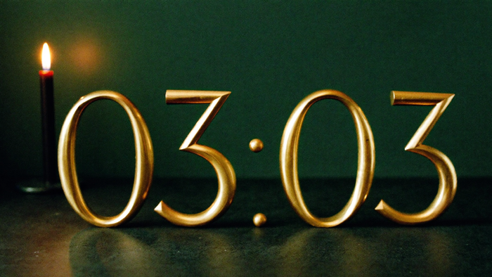 Brass numerals reading 03:03 on a dark stone ledge in front of a deep green wall, a lit candle on the left