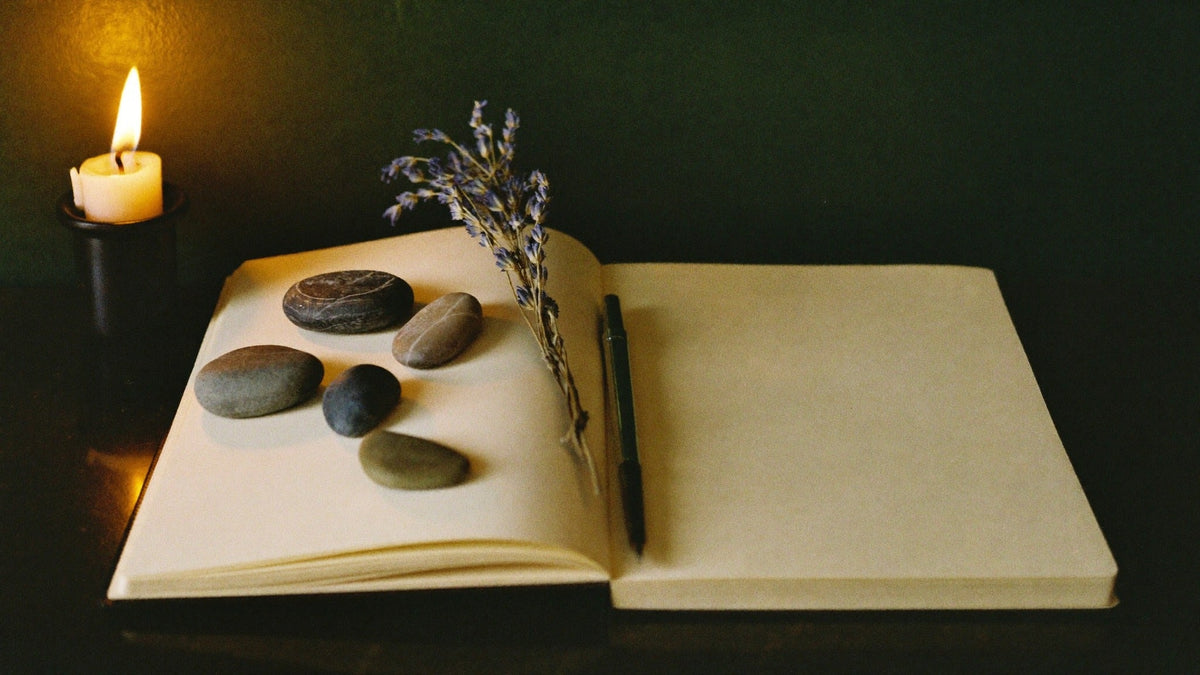Open blank notebook with a black pen, five smooth river stones and a sprig of dried lavender beside a lit candle