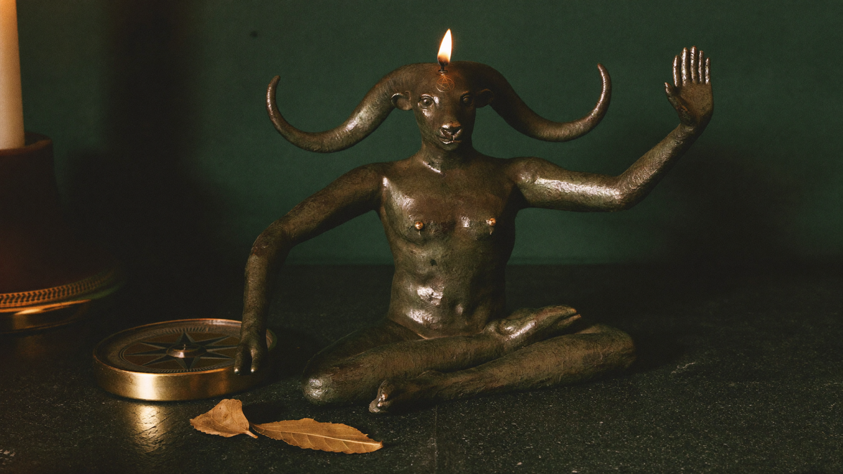 Bronze goat-headed figure seated cross-legged, one hand raised, a lit candle burning above its horns